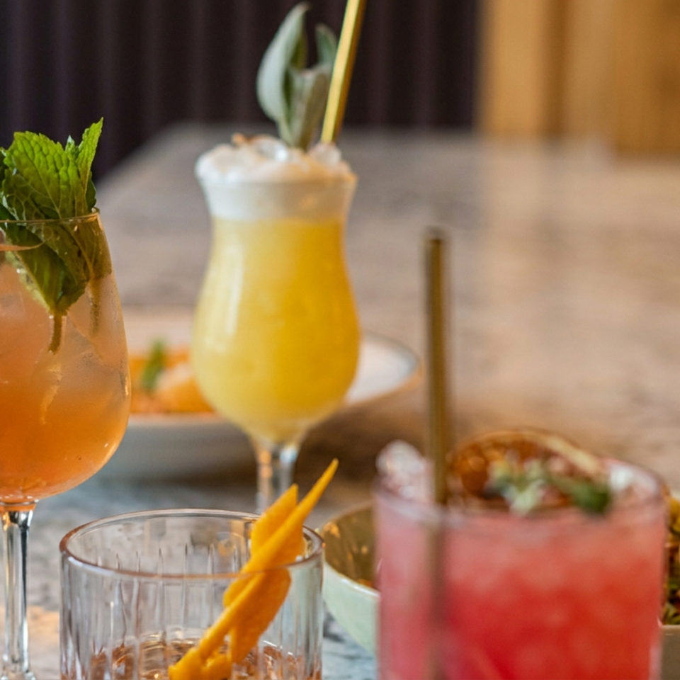 Colorful cocktails with mint, citrus garnishes, and straws arranged on a restaurant table beside bowls of food.