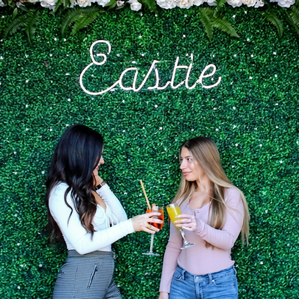 Two women toast with colorful drinks in front of a greenery wall decorated with flowers and the word “Gastro.”